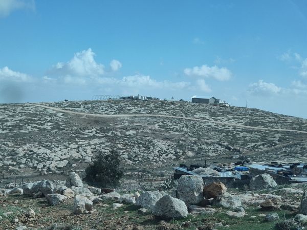 Utsikt från huset, närmast i bilden ser man ett par stenar och ett par små hus, där bor djuren. På en höjd syns ett par större hus och ett stort växxthus, tillhörande israeliska bosättare.